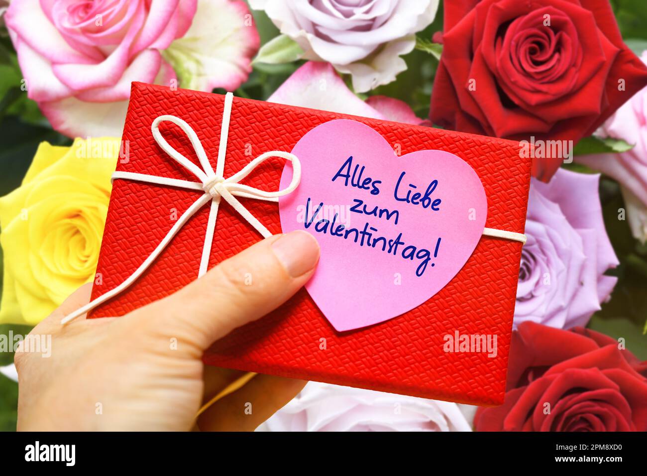 FOTOMONTAGE, Hand hält Geschenk mit einem herzförmigen Zettel und der Aufschrift alles Liebe zum Valentinstag vor einem Blumenstrauß Stock Photo