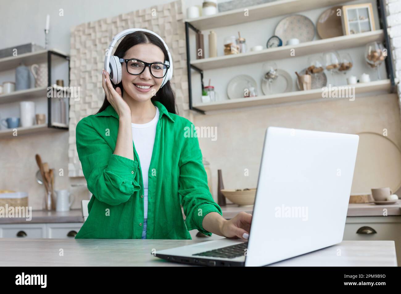 A young woman in headphones sits at home in the kitchen in front 
