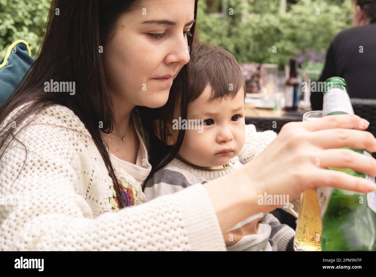 A new mother is pouring a nonalcoholic beer in her glass while holding her infant sitting in a