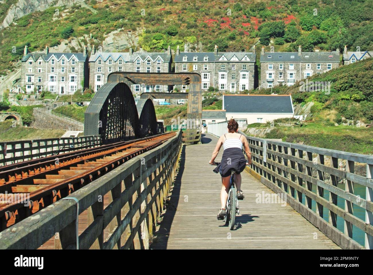 Barmouth Gwynedd in North Wales a row of similar blocks of homes including Guest Houses & other holiday accommodation with stunning views over this Afon Mawddach river estuary with easy access to walks and bike rides along this Victorian railway bridge footpath towards the Snowdonia National Park on the opposite shoreline.Back view of young woman cyclist arriving from Morfa Mawddach on the same opposite shore. Stock Photo