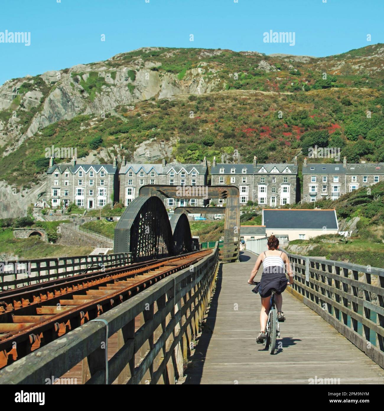 Woman cyclist completes a bike ride journey beside the historical Victorian Barmouth Bridge (or Pont Abermaw) or (Barmouth Viaduct) which is a Grade II listed single track wooden railway structure. Parallel to the train track is this footbridge walkway & both cross the estuary of the Afon Mawddach River linking Barmouth here here to Morfa Mawddach a small station 820 metres  away both in Gwynedd in North Wales. The footbridge is part of the National Cycle Route 8 & is available to pedestrians cyclists and motorbike users. Stock Photo