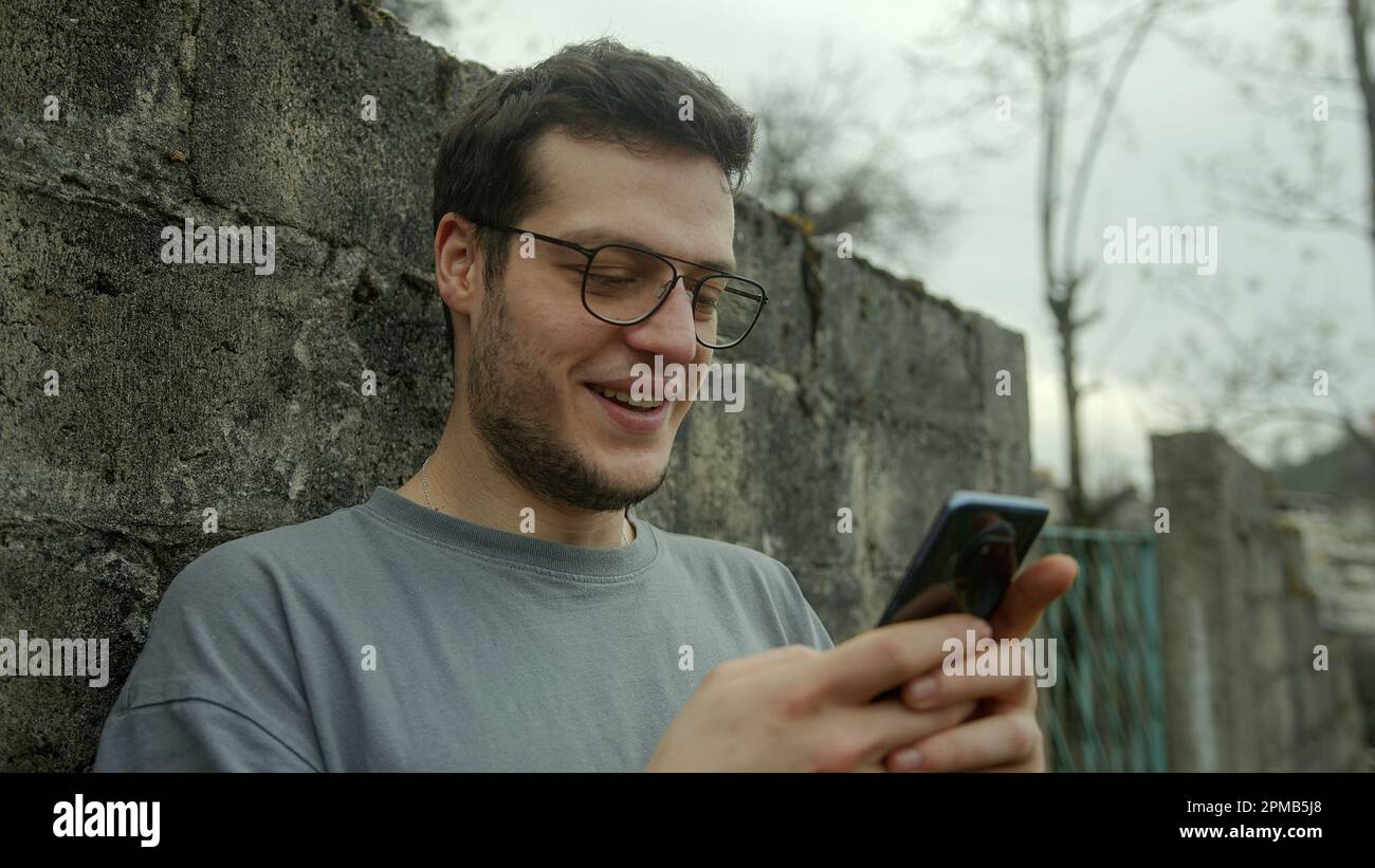 Handsome Young Man with Glasses Surprised and Smiling While Using Smartphone Outdoors Stock Photo
