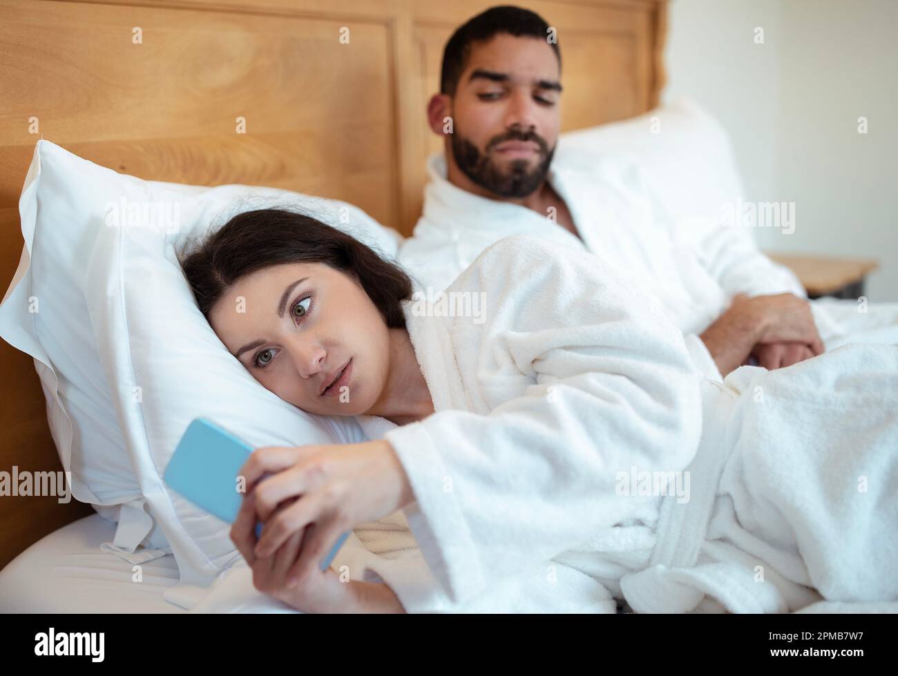 Husband Looking At Cheating Wife Texting On Smartphone In Bedroom Stock