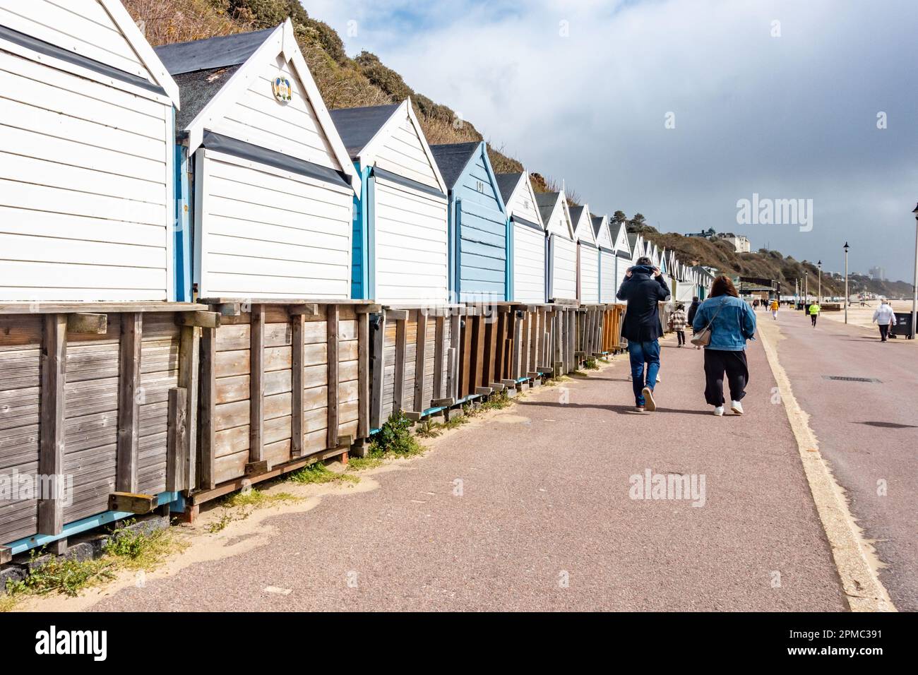 People walk along a path in front of beach huts on Bournemouth Beach in Dorset, UK under a stormy, grey sky Stock Photo