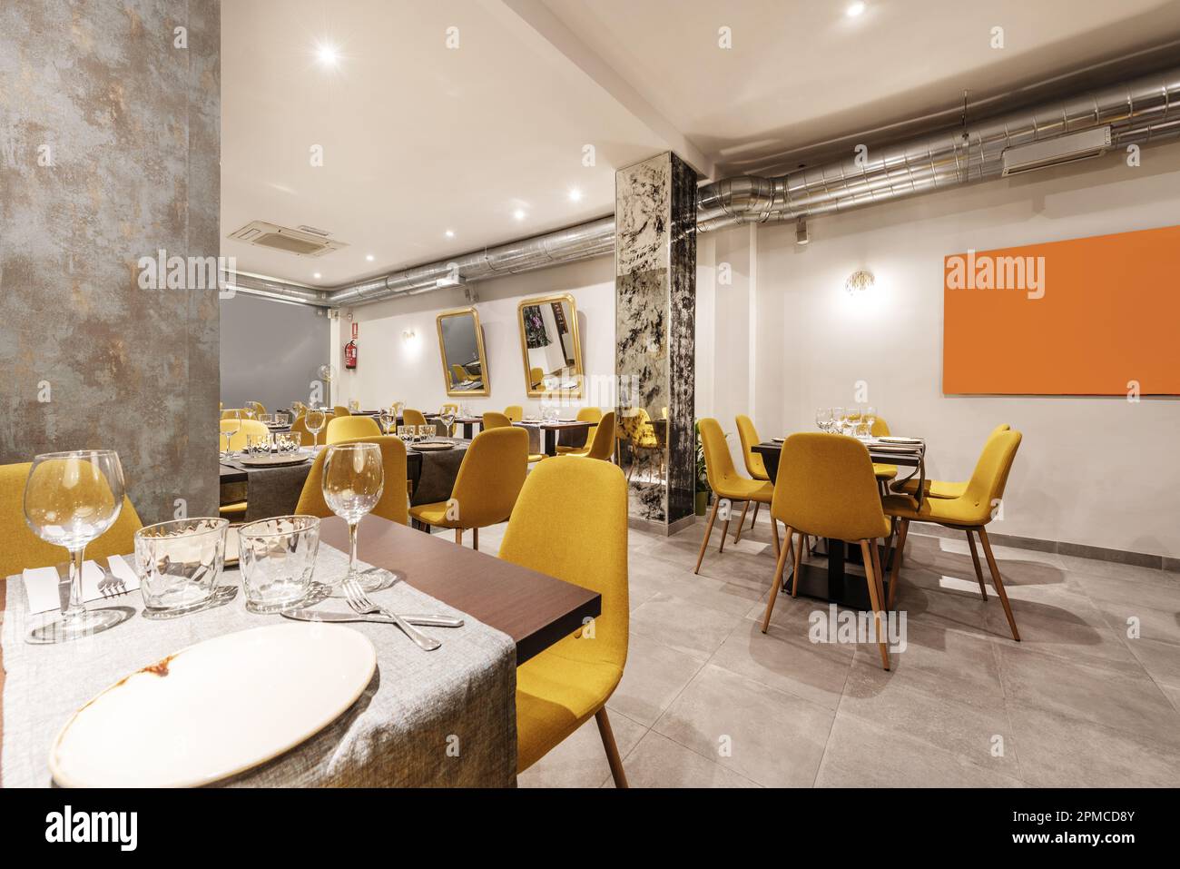 Dining room of a restaurant with tables, with cutlery, crockery, glassware and assembled chairs upholstered in yellow fabric and exposed air condition Stock Photo