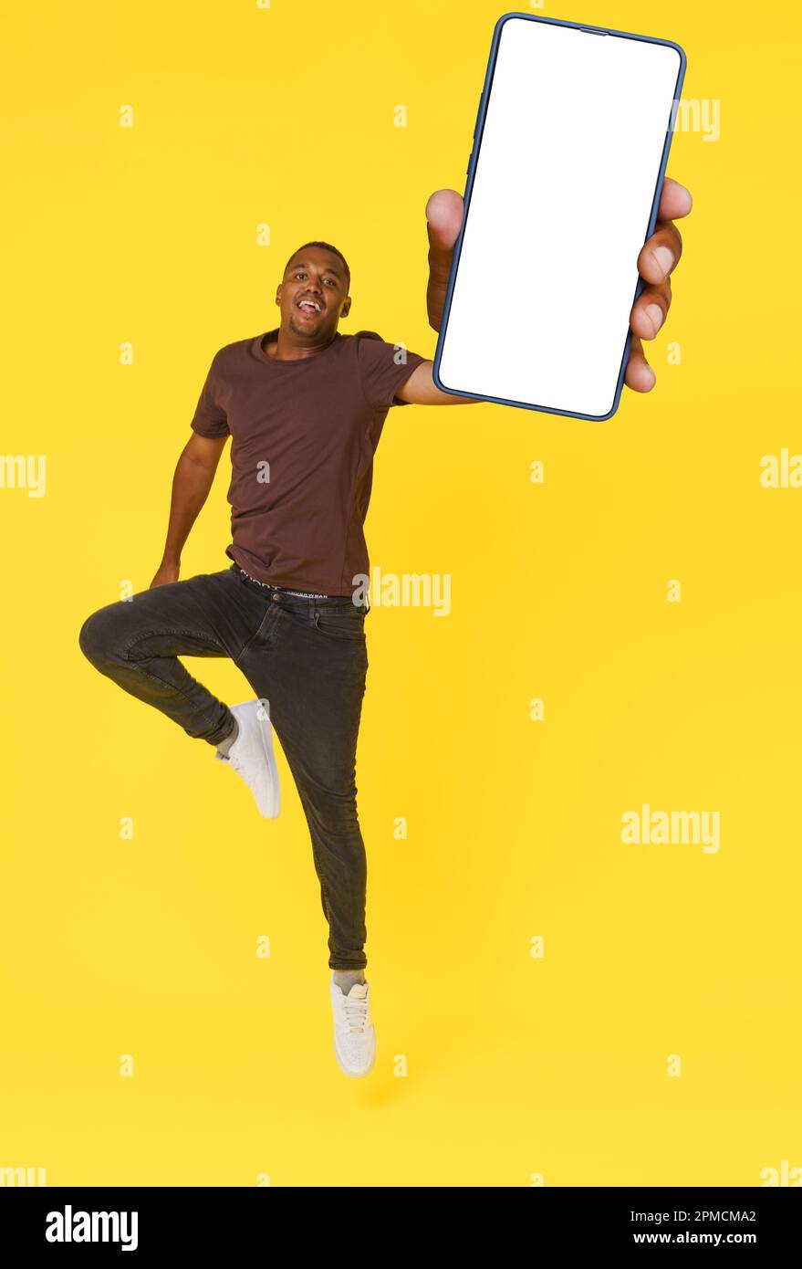 Energetic African American student is captured jumping with mobile phone featuring empty white screen in hand, on yellow background. This image is perfect for product placement or advertising concepts. High quality photo Stock Photo