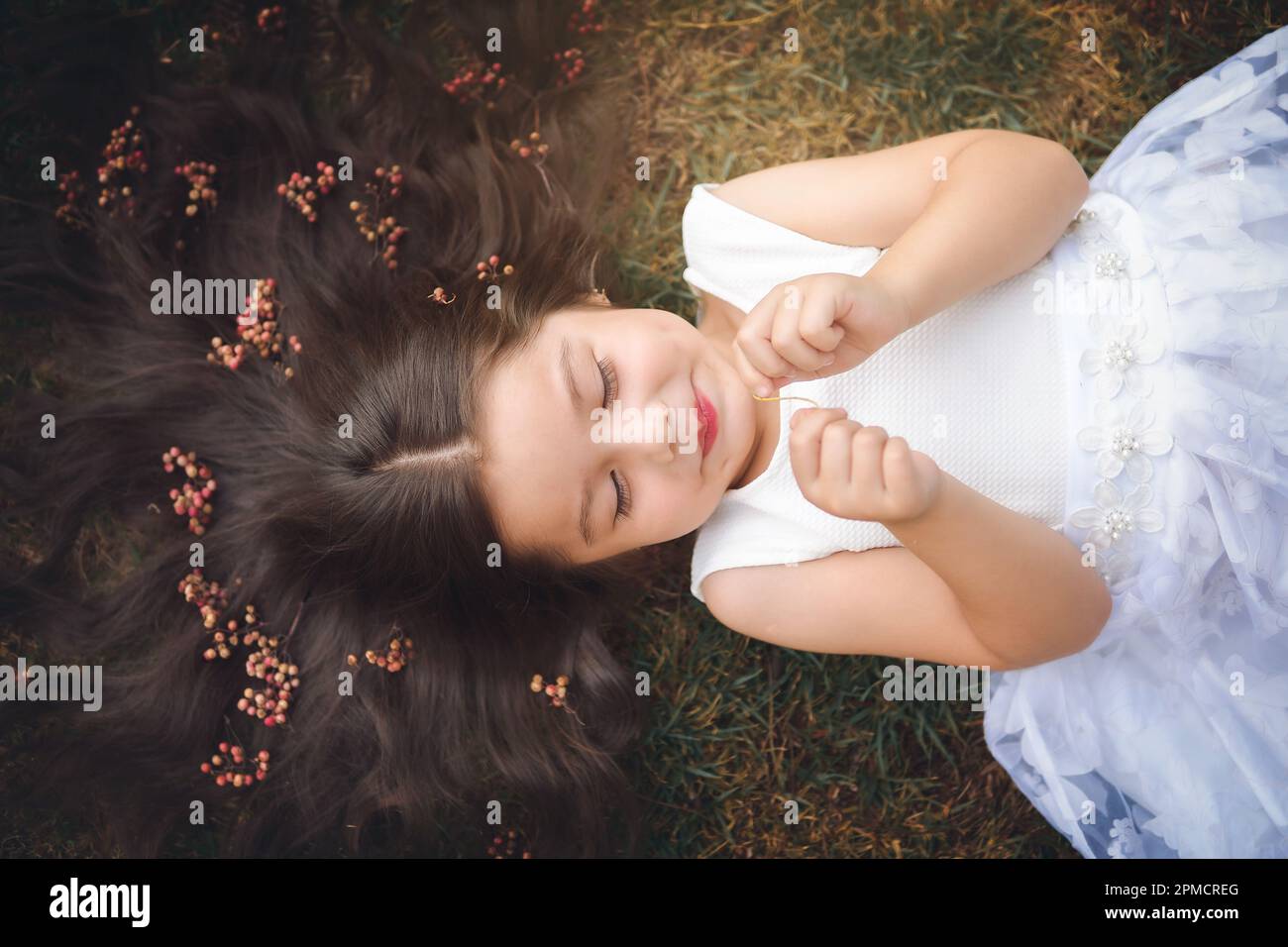 Little girl in white dress lying on the grass, she is smiling, her hair is very long and princess-like, she plays with her hands on the theme of child Stock Photo