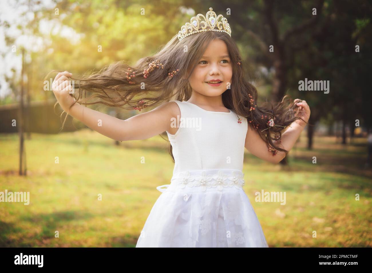 Little girl in white dress and princess crown, she plays with her long blonde hair, copy space, children's day theme. Stock Photo