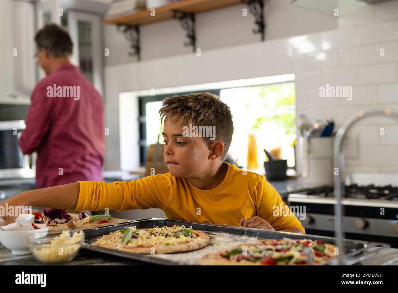 Cute caucasian boy preparing pizzas in tray over kitchen island with grandfather in background Stock Photo