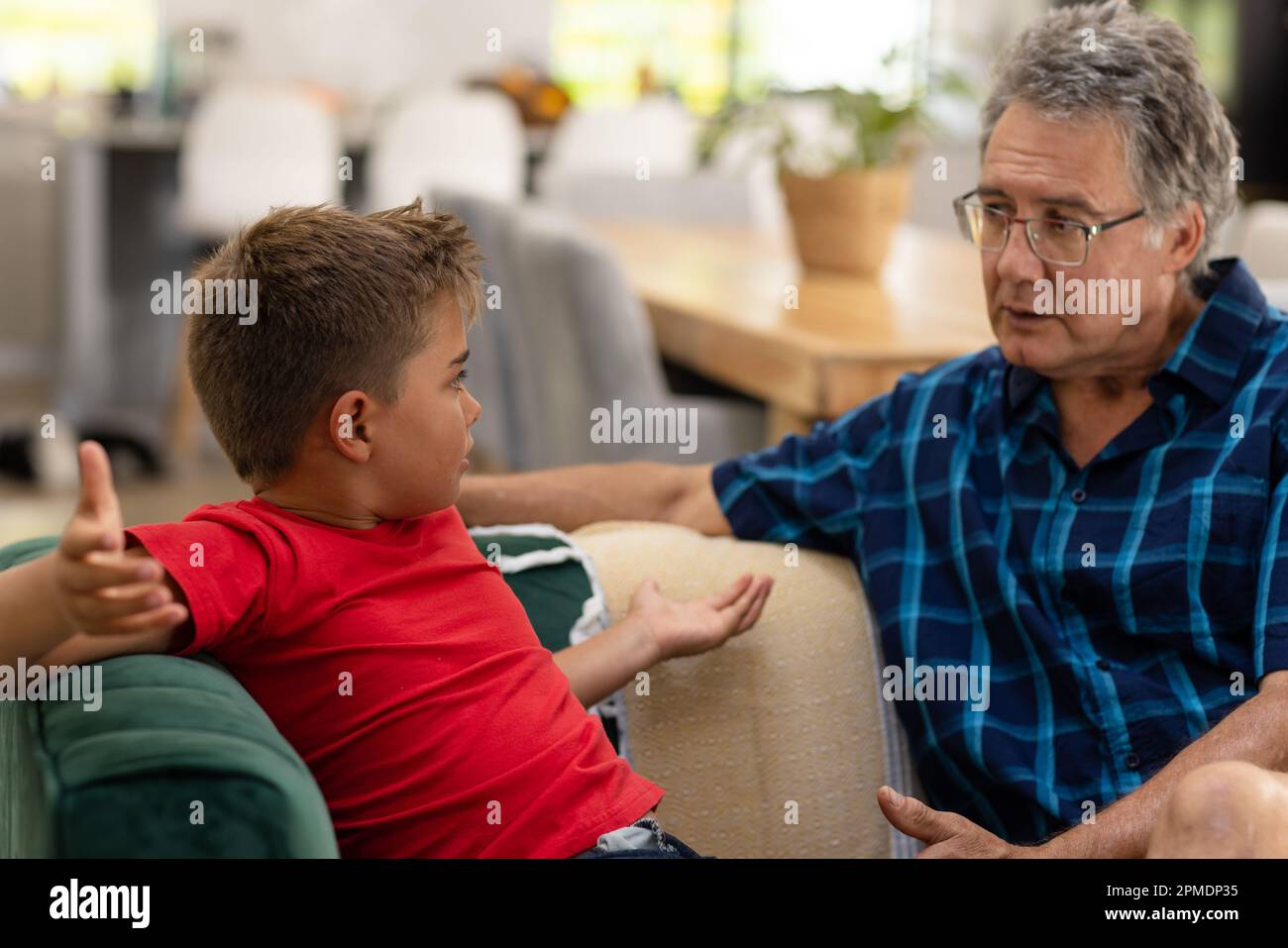Caucasian angry boy shrugging shoulders while talking with grandfather on sofa in living room Stock Photo