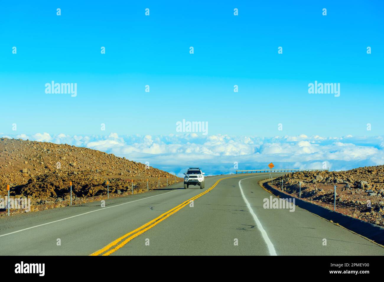 A breathtaking landscape of a winding road along a cliff with a railing, high above the clouds, and a car approaching towards the viewer. Stock Photo