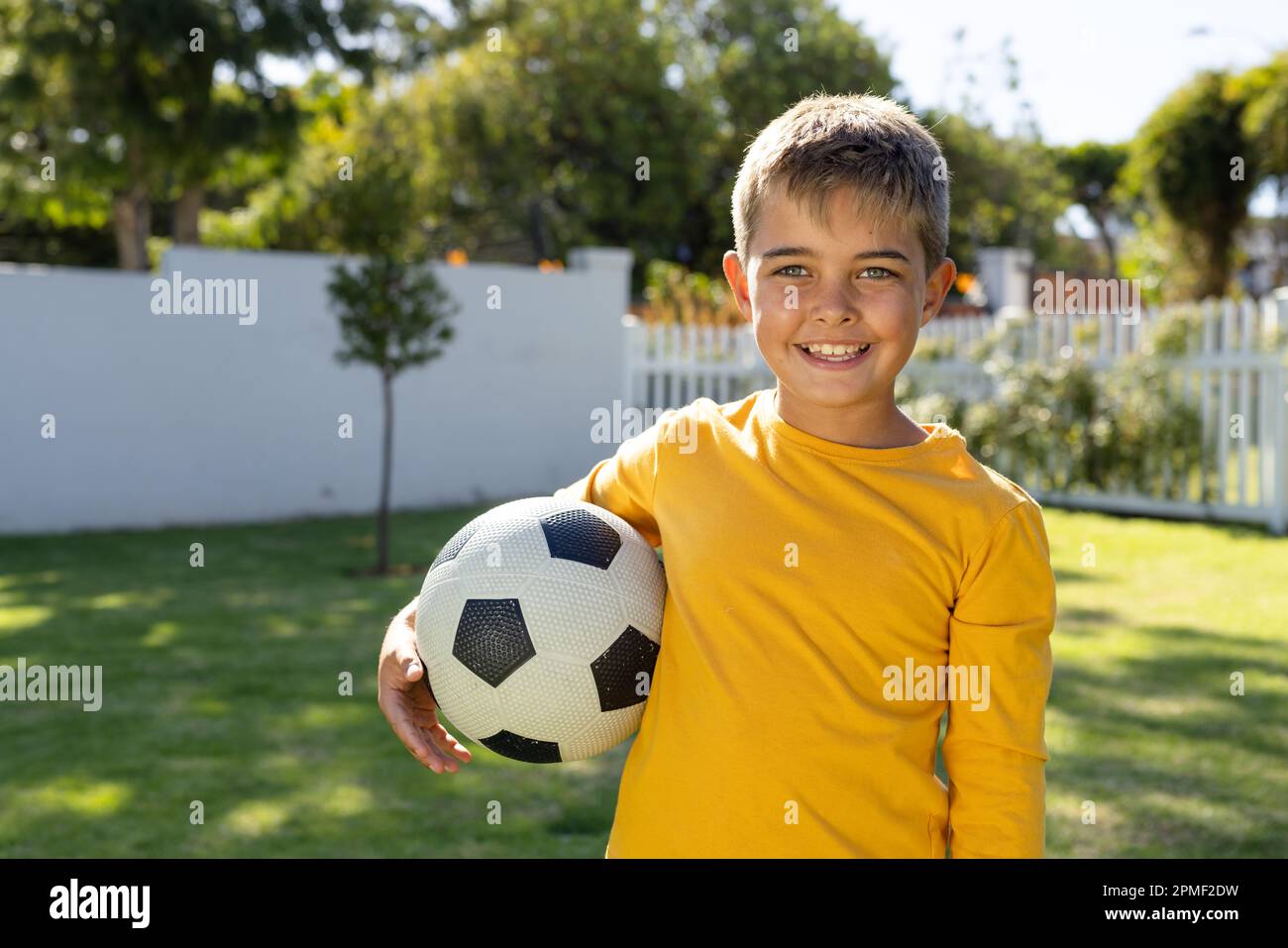 Portrait of cute caucasian boy smiling and holding soccer ball while standing on grassy land in yard Stock Photo