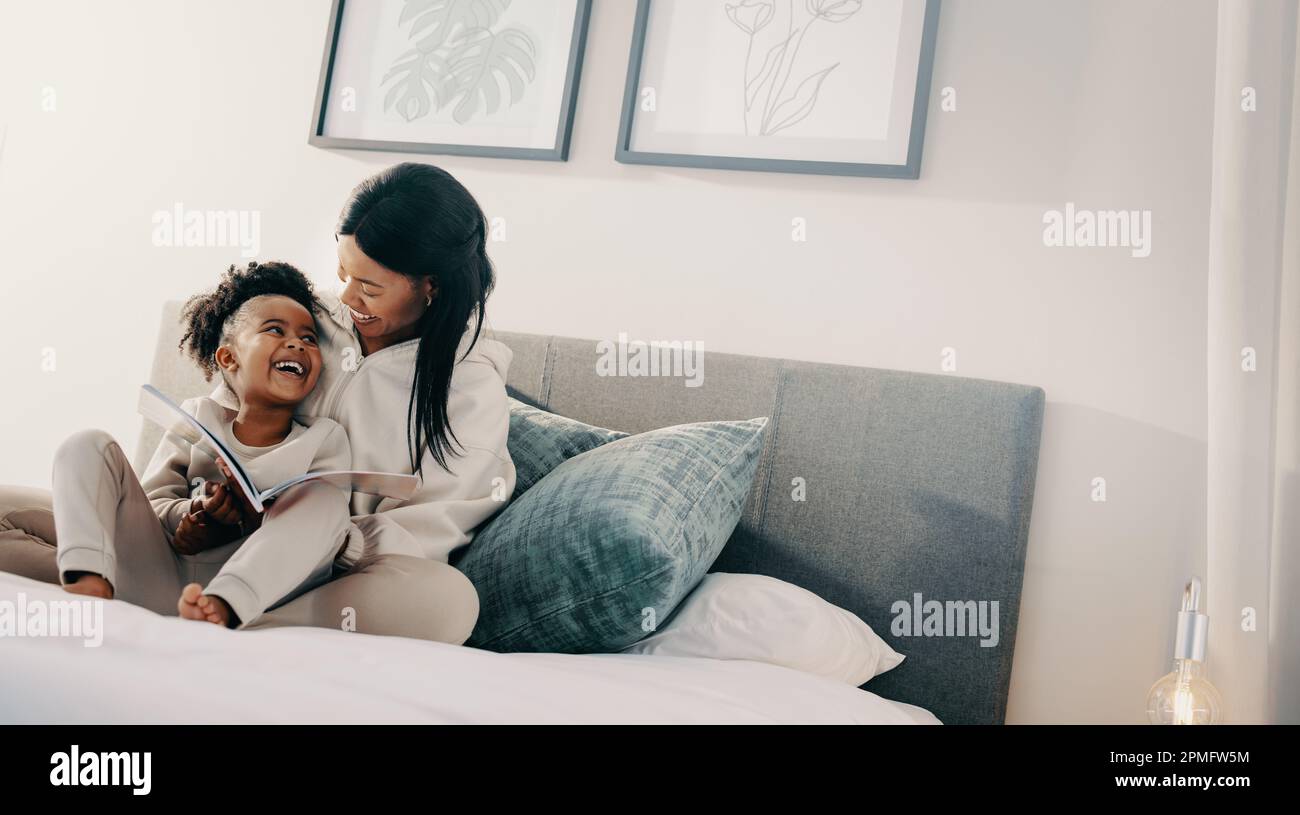 Woman smiles at her child while helping her to read a book, this mother and daughter are sitting on a bed. Single motherhood and parental love. Stock Photo