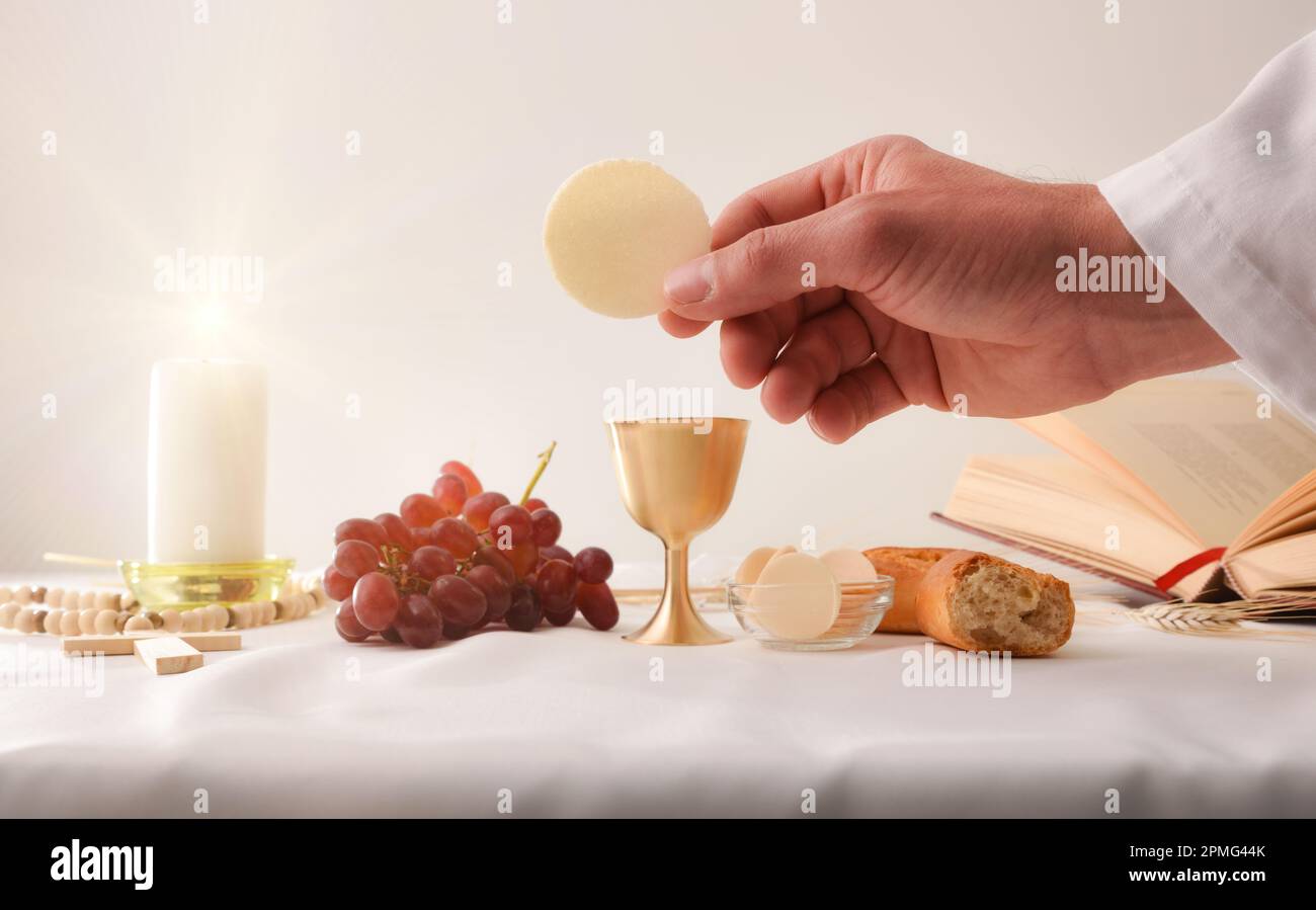 Hands of a priest consecrating a host as the body of Christ to distribute it to the communicants with table with sacred objects behind. Stock Photo