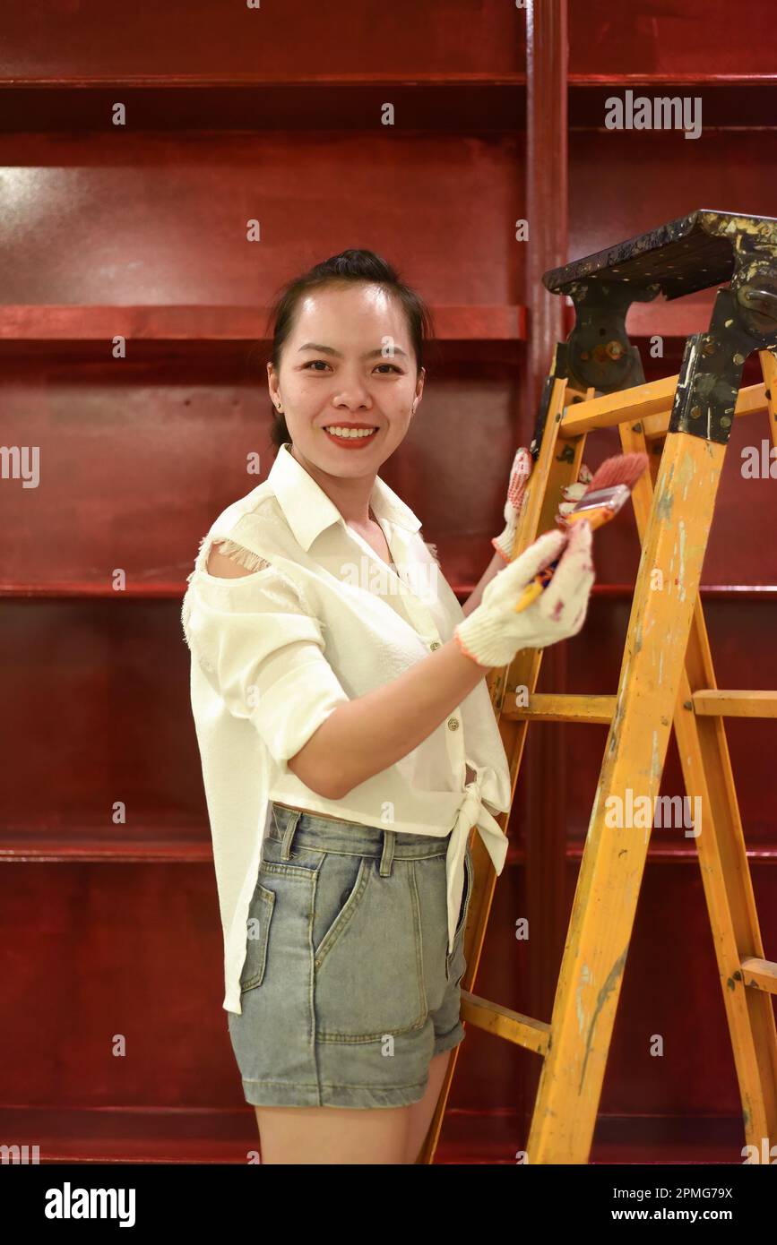 Vietnamese asian woman stands on a stepladder with a brush in her hand and paints the wall, makes repairs Stock Photo