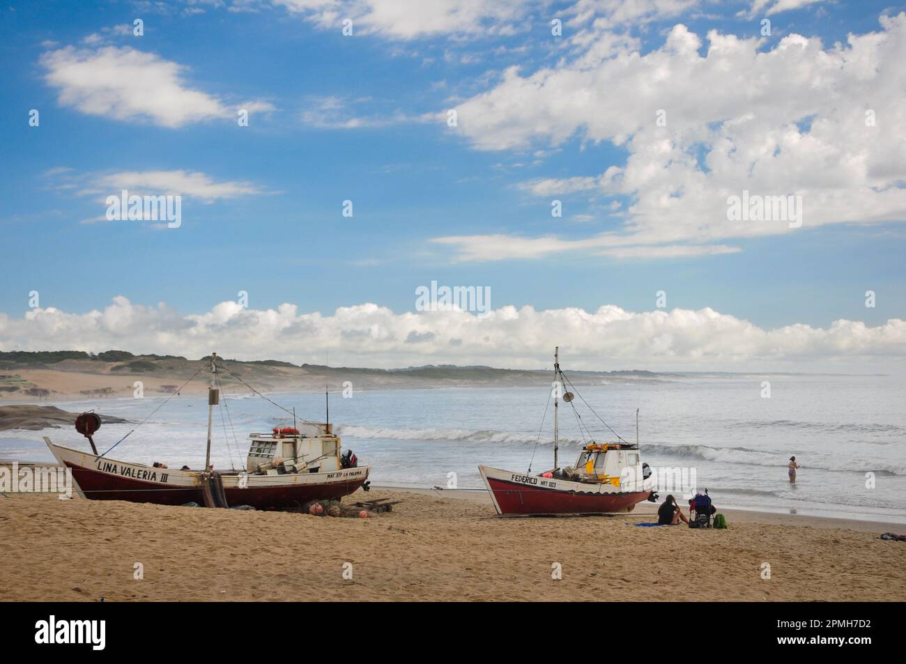 A couple of fishing boats on the sand of Punta del Diablo beach, while one person rests on the sand and another swims in the sea Stock Photo