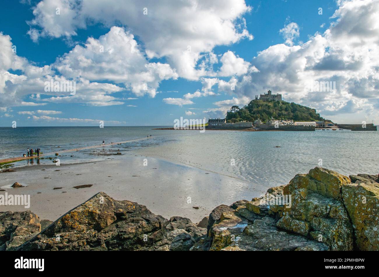 St Michaels Mount off the coast of Cornwall near Marazion on a clear sunny day in April. Rocks in the foreground and people walking across the sea. Stock Photo