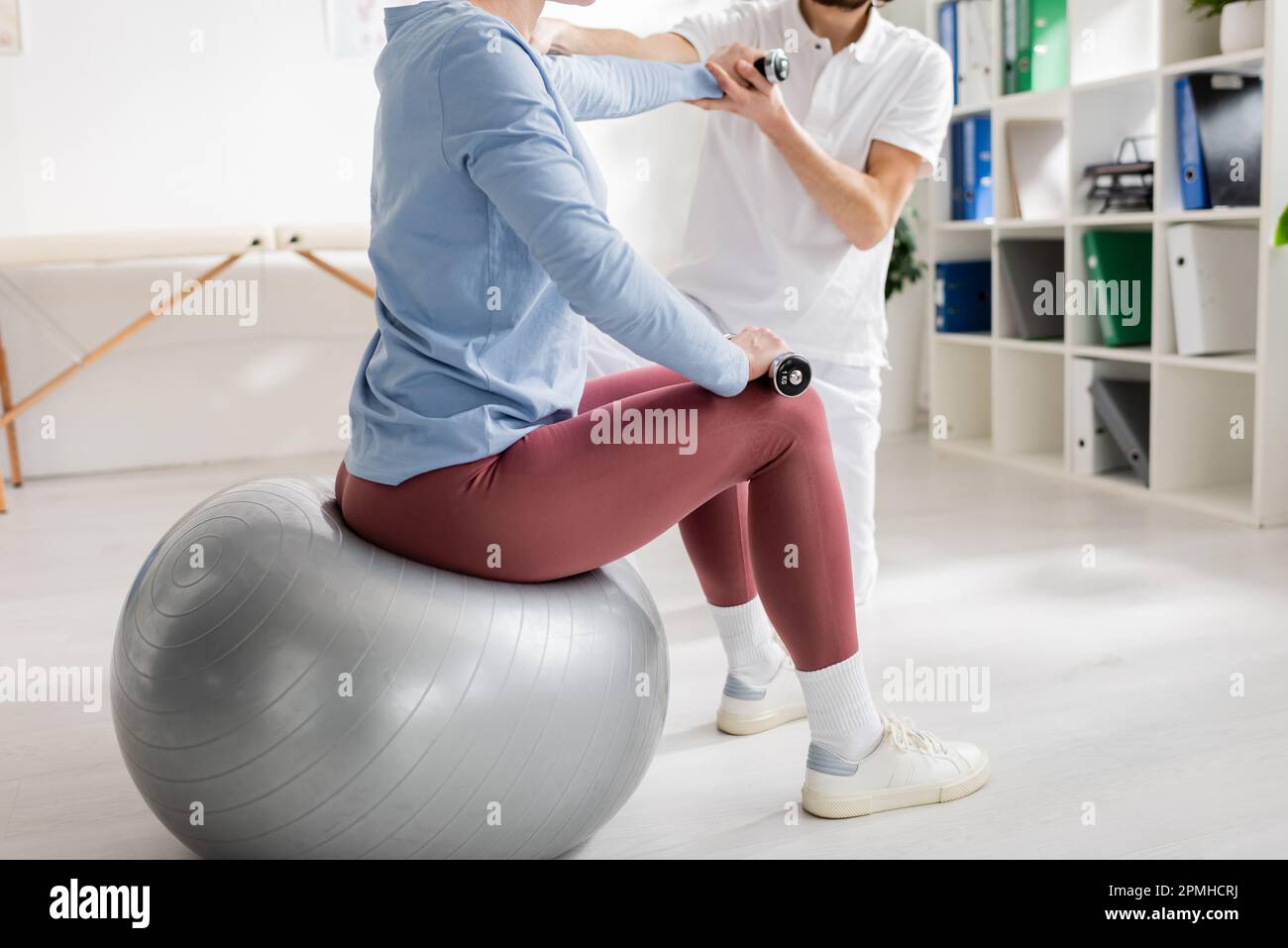 cropped view of woman sitting on fitball and exercising with dumbbells near physiotherapist in rehabilitation center,stock image Stock Photo