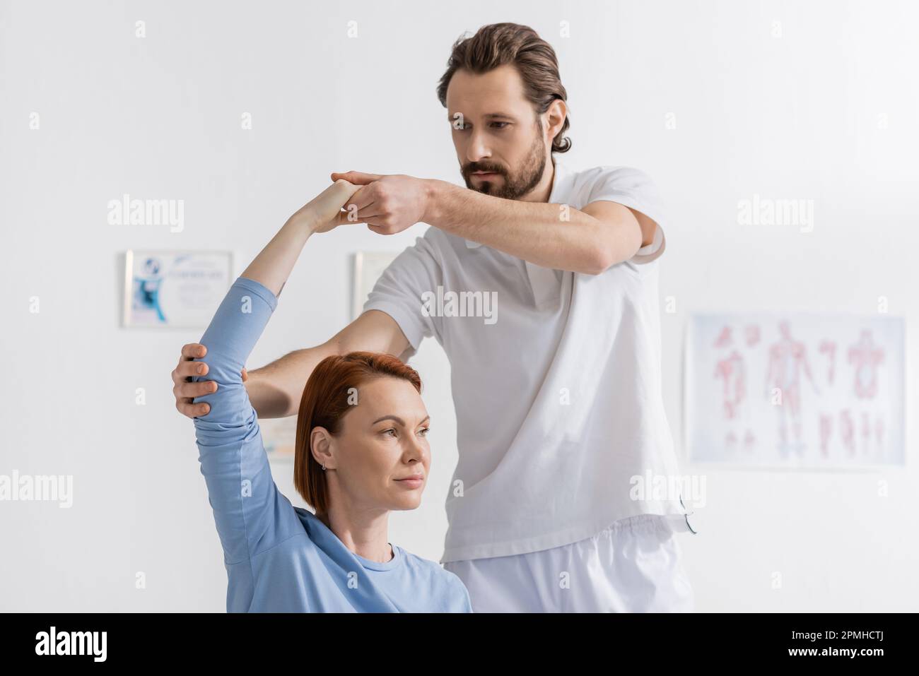 bearded physiotherapist supporting arm of redhead woman exercising in rehabilitation center,stock image Stock Photo