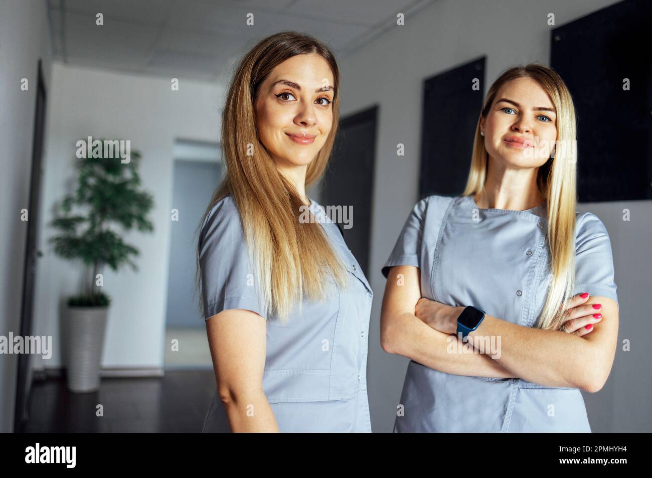 Beautiful smiling female doctors in medical uniform stands near the window in clinic. Young nurse is talking. Sunny day. Light interior. Copy space. Stock Photo