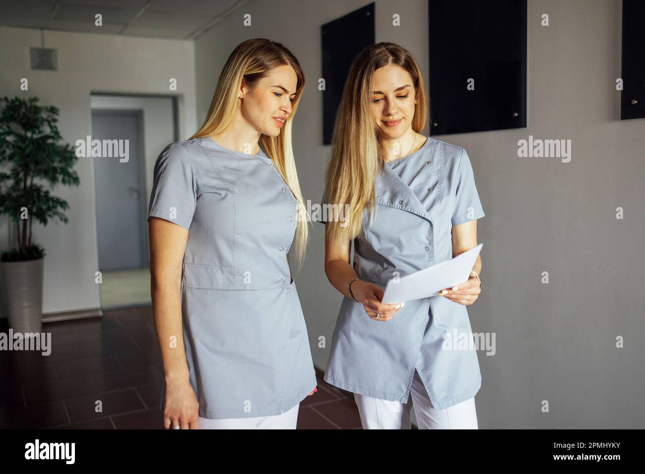 Beautiful smiling female doctors in medical uniform stands near the window in clinic. Young nurses is holding sheets of paper in her hands. Sunny day. Stock Photo