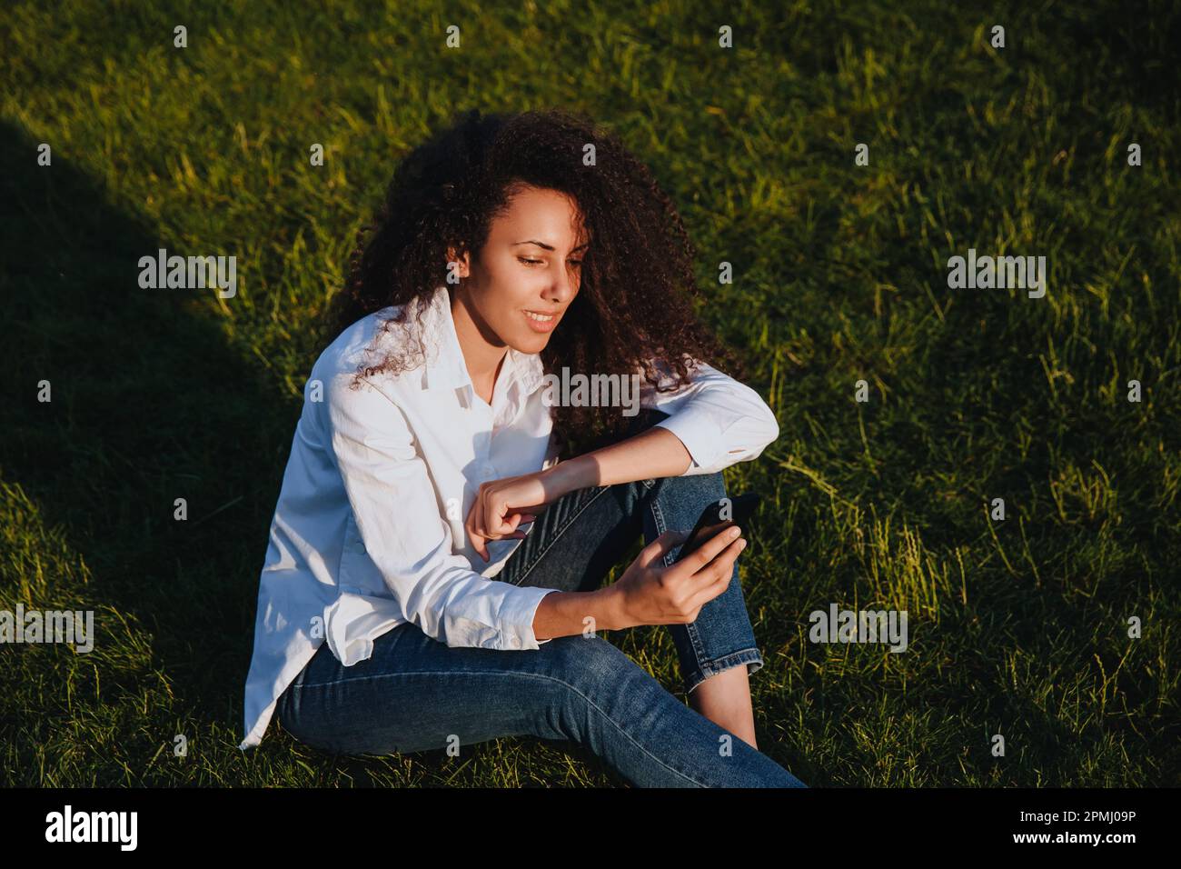 A young girl sits on the green grass in the park and uses social networks using a mobile phone. Stock Photo