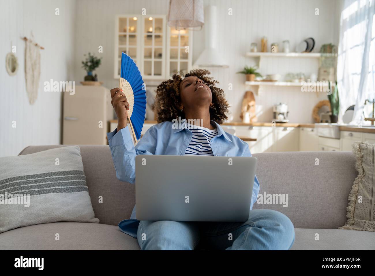 Exhausted overheated African American girl freelancer waving hand fan and working on laptop at