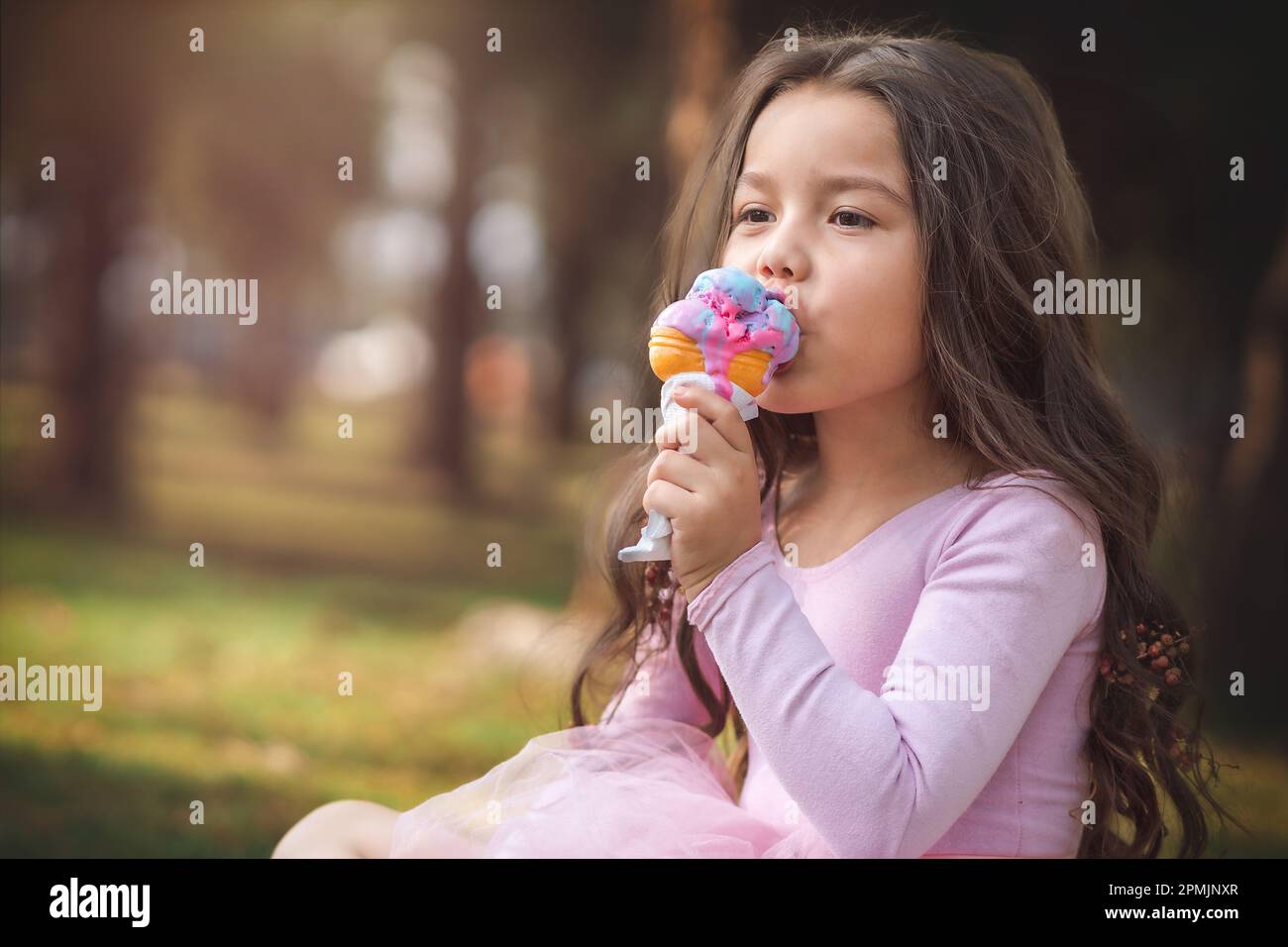 Cute blonde curly haired girl eating a nice ice cream in the forest on a nice summer day, children's day concept, happy girl enjoying a hot day. Stock Photo