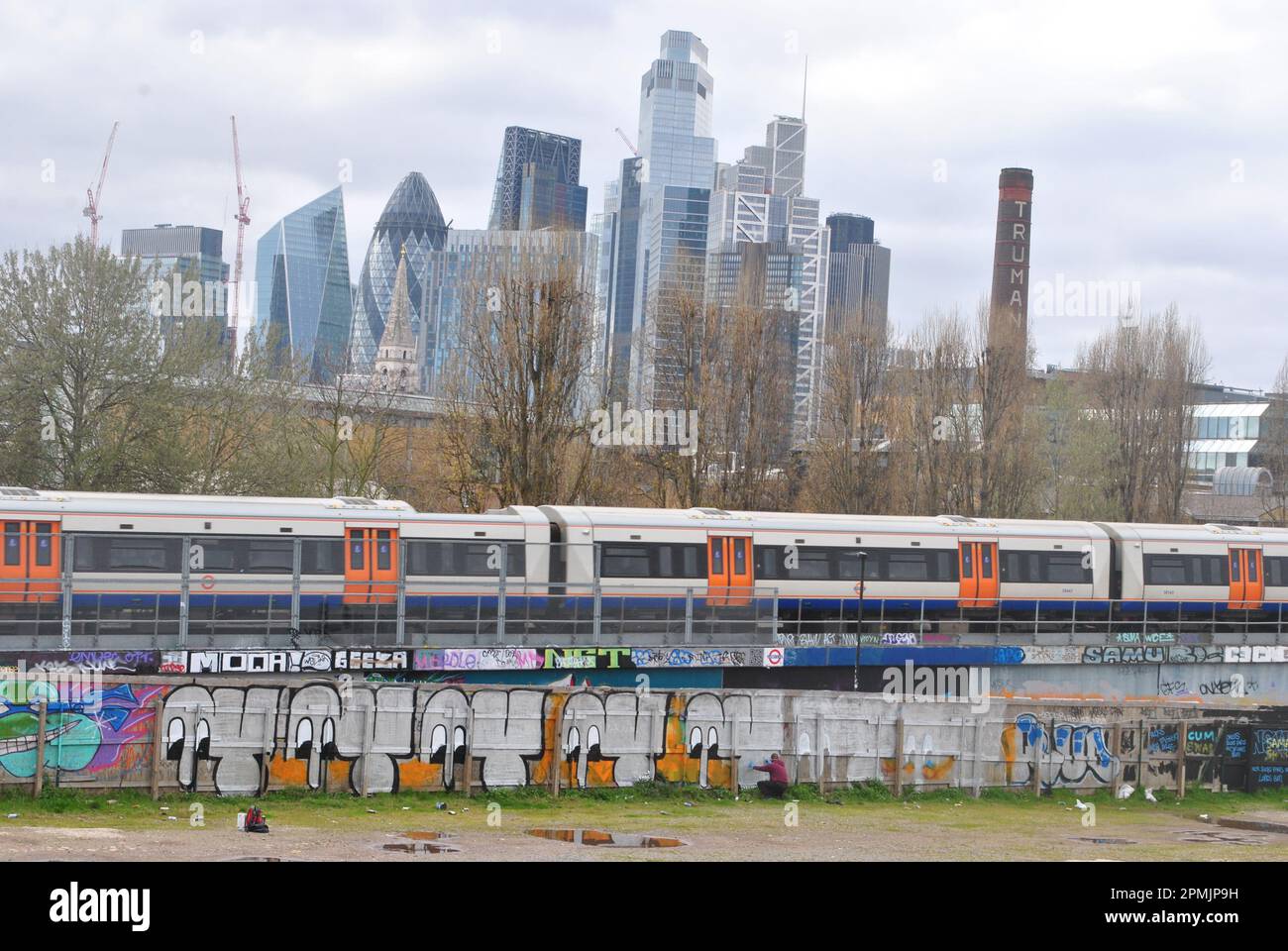 A GRAFFITI ARTIST PAINTS IN SHOREDITCH, EAST LONDON WITH A PASSING OVERGROUND TRAIN AND A VIEW OF THE CITY OF LONDON BEYOND HIM. Stock Photo