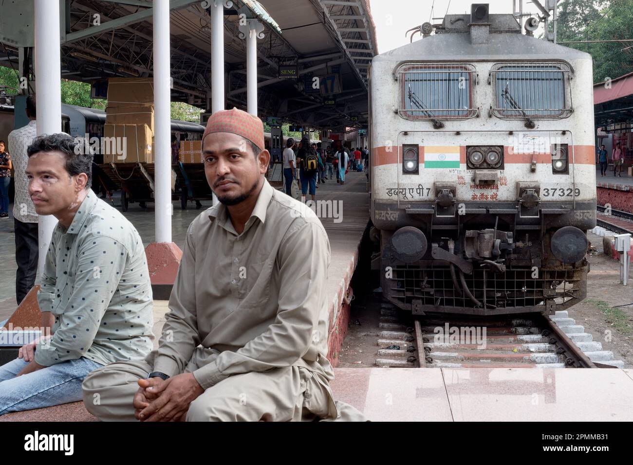 Passengers sitting on a platform at Chhatrapati Shivaji Maharaj Terminus in Mumbai, India, with a WAP-7 type diesel-electric locomotive behind them Stock Photo