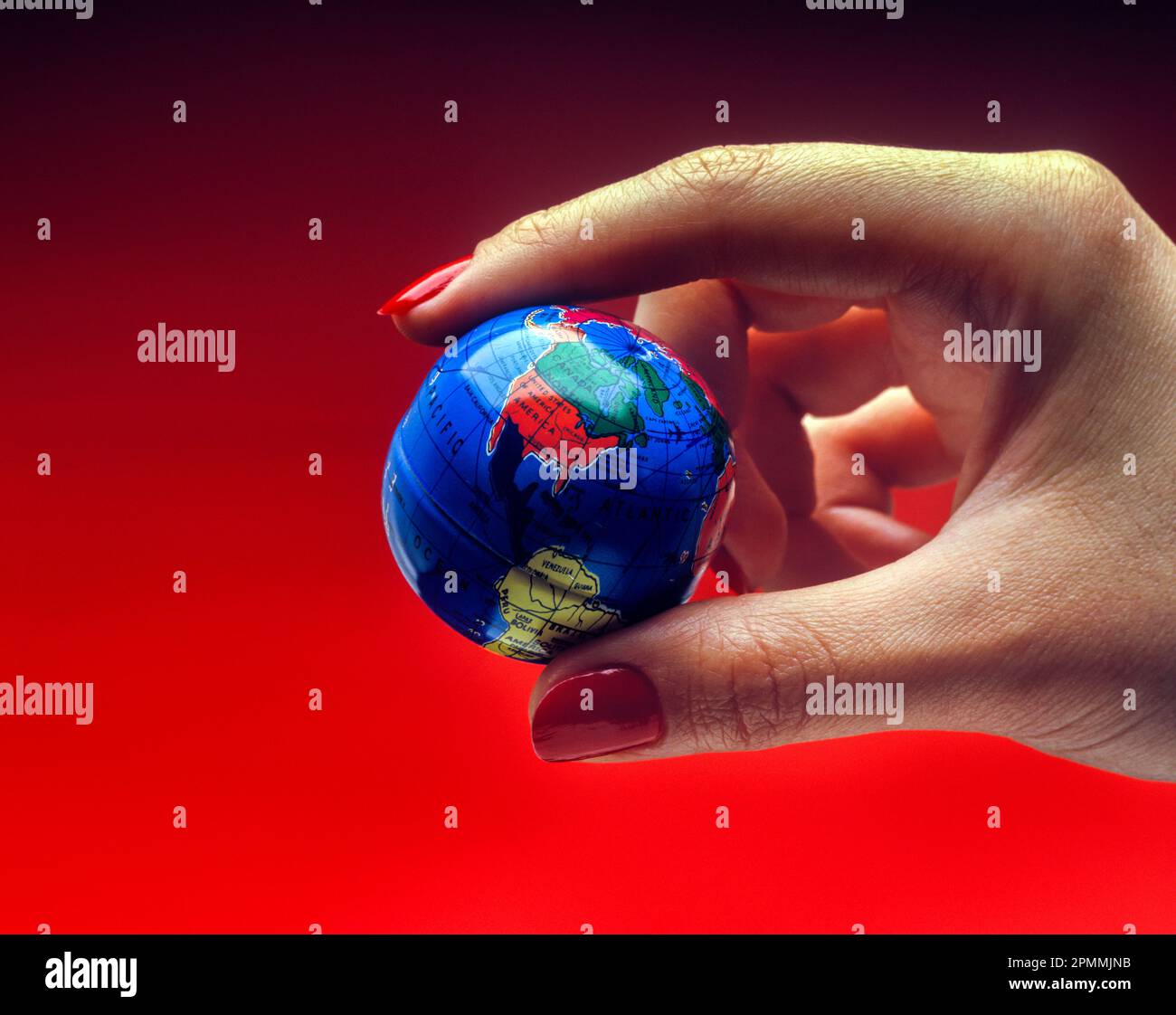 1991 HISTORICAL WOMAN’S HAND HOLDING TOY PLANET EARTH GLOBE ON RED BACKGROUND Stock Photo