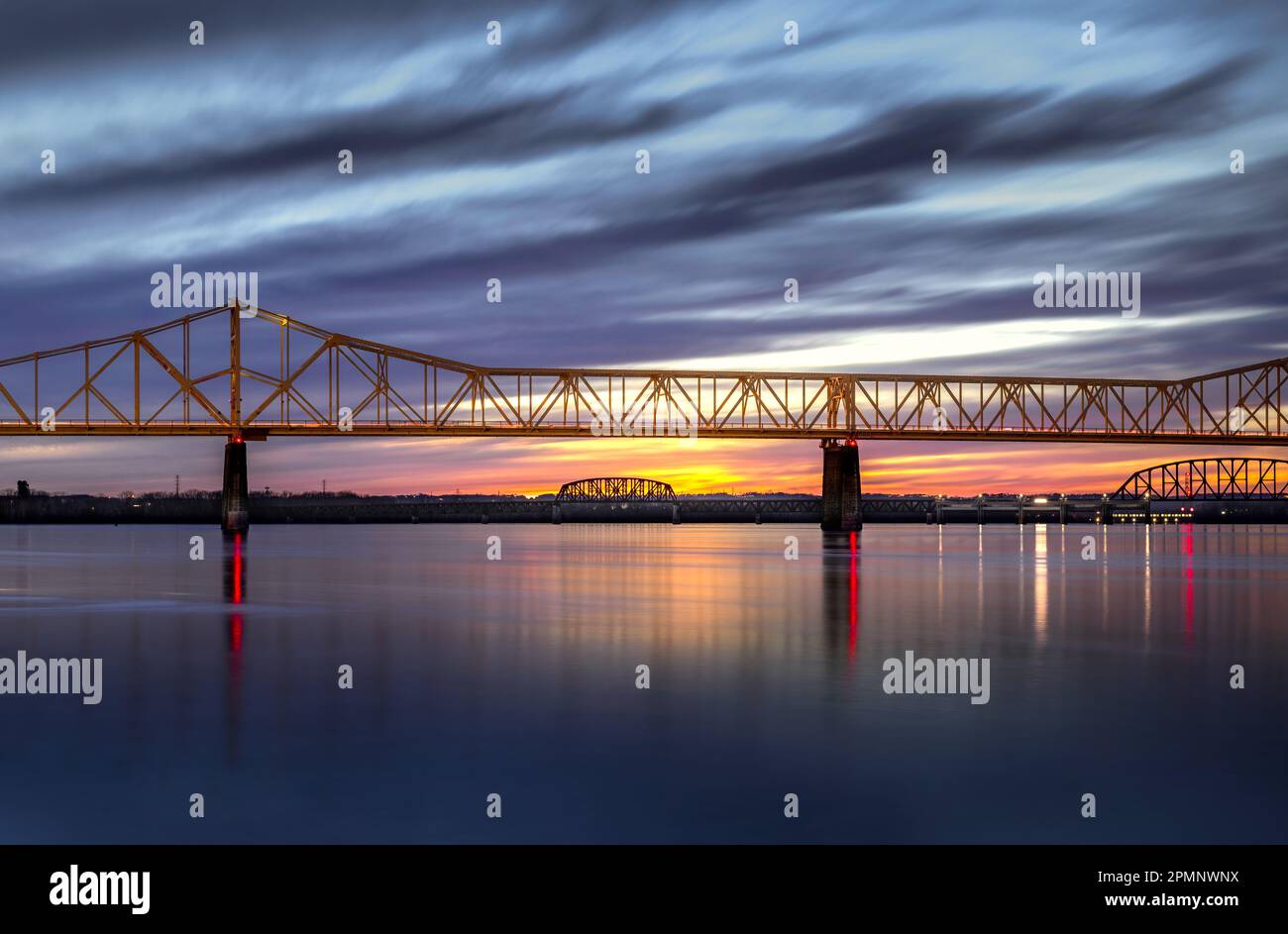 A stunning landscape featuring a bridge spanning over a tranquil body of water, illuminated by a beautiful sunrise, with clouds in the background Stock Photo
