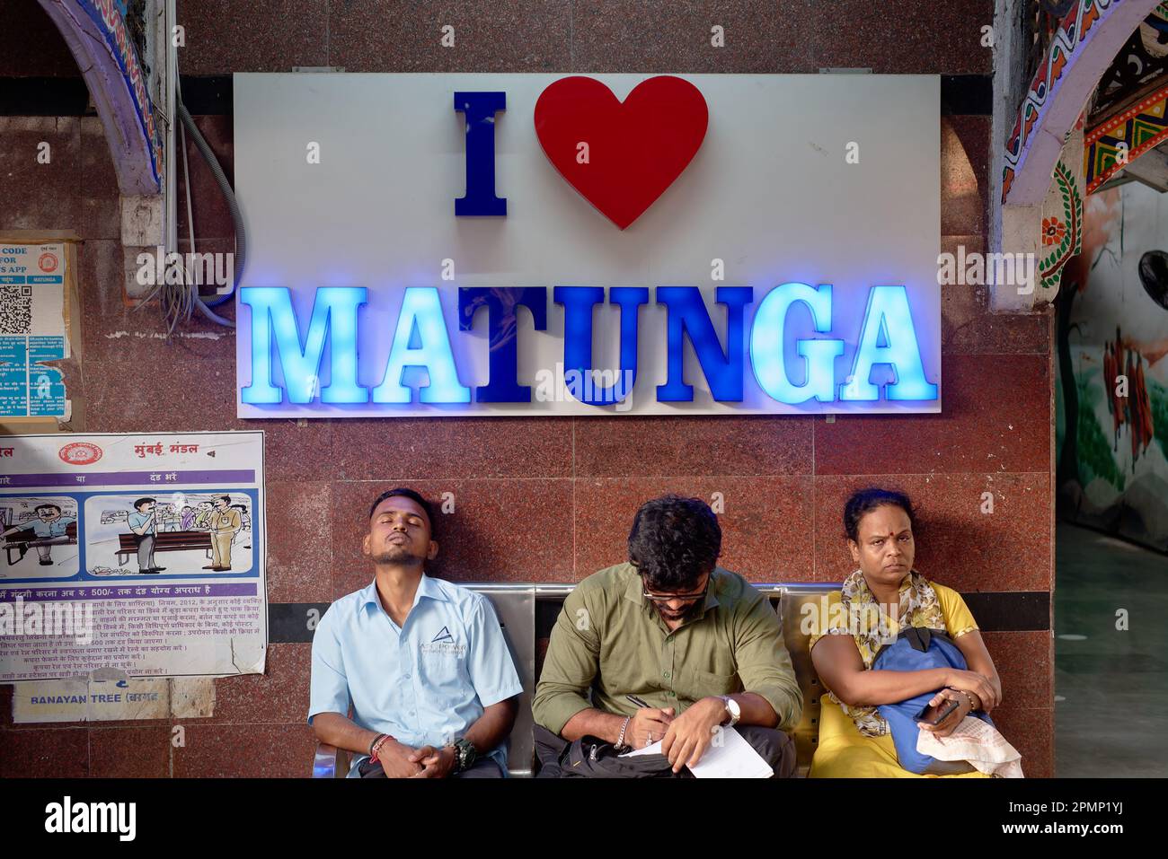 Three passengers sit on a bench at Matunga railway station (Central Railways) in Mumbai, India, under an illuminated neon sign saying 'I Love Matunga' Stock Photo