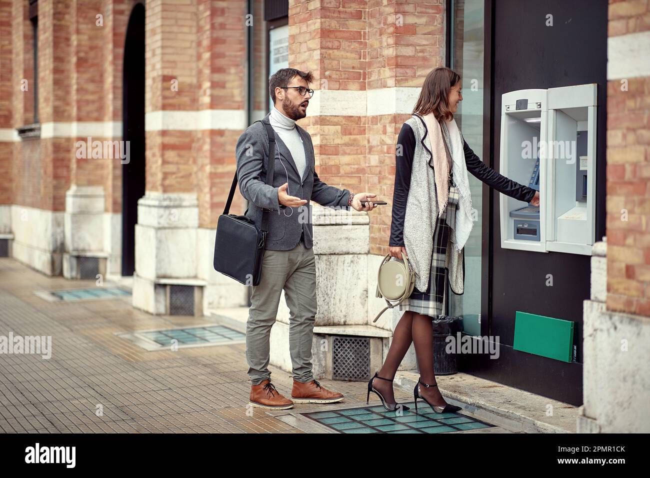 Impatient man protest in front cash machine while woman takes her money Stock Photo