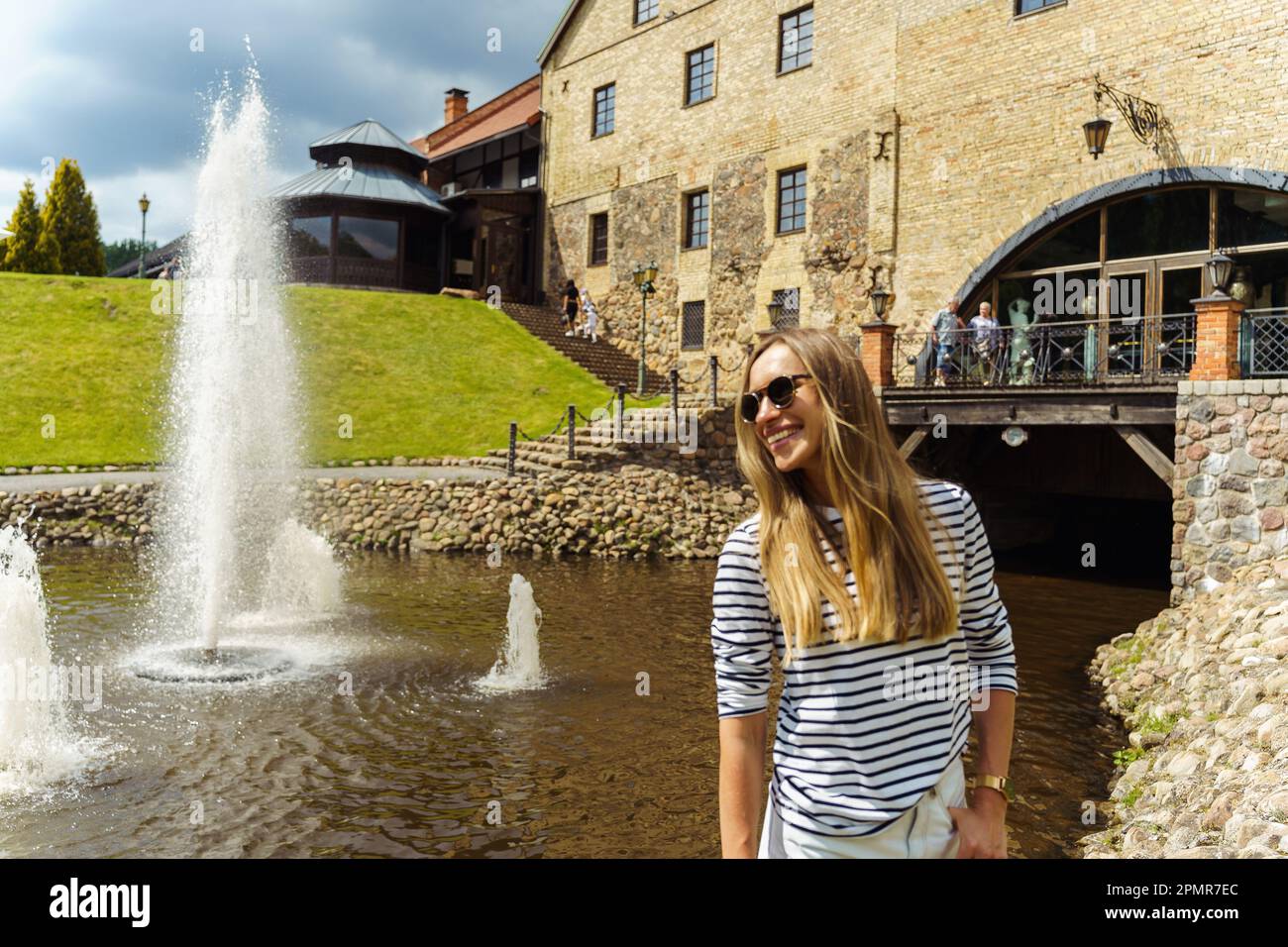 A long-haired woman in a striped jumper and sunglasses laughs next to a fountain in the summer. Stock Photo