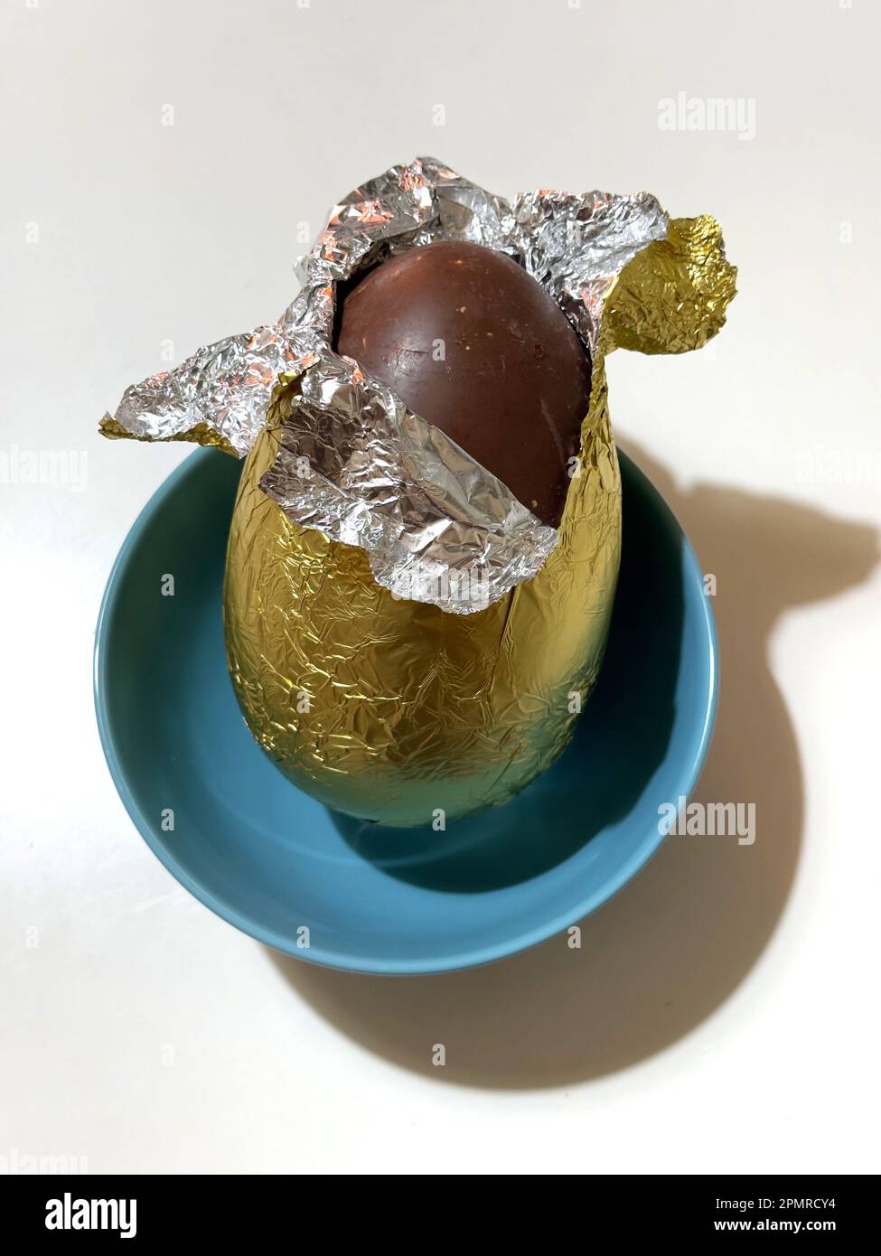 a chocolate easter egg, wrapped in tin-foil, in a turquoise bowl against a white background Stock Photo