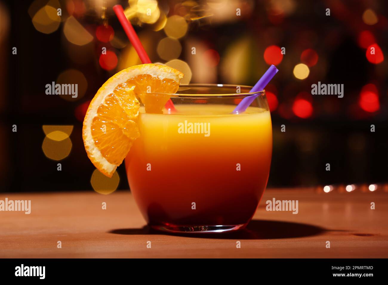 Glass of tasty Tequila Sunrise with orange slice on table against blurred lights Stock Photo