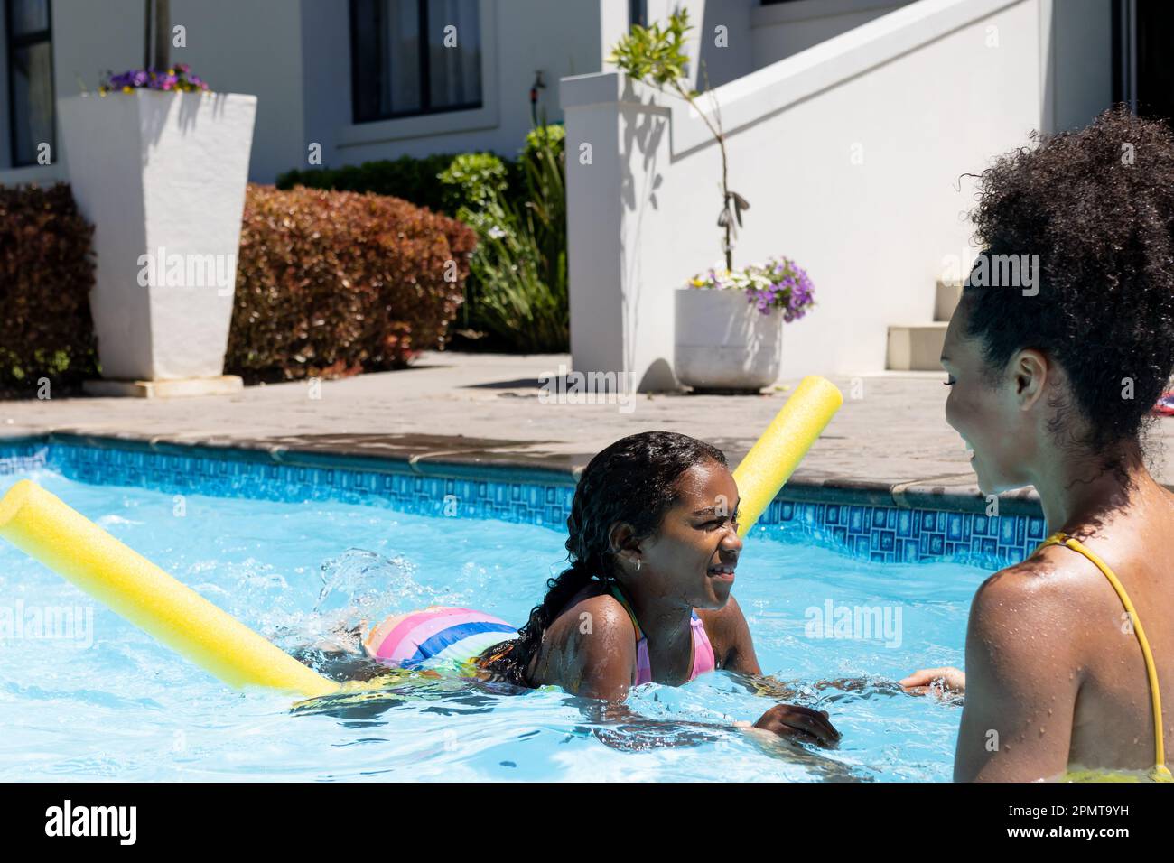 Happy biracial mother and daughter learning to swim with float in swimming pool in sunny garden