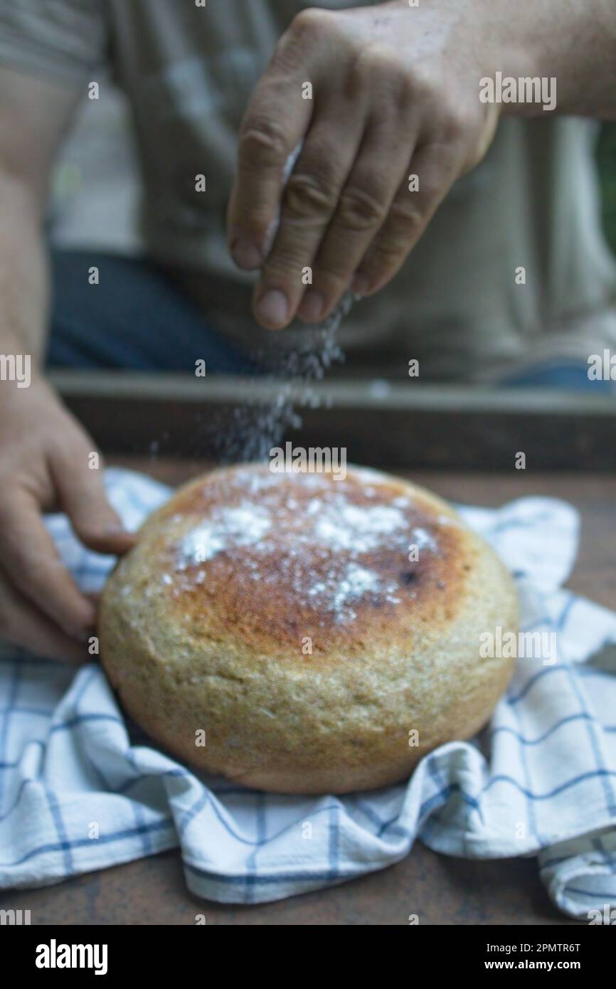 Baker's hand sprinkles flour on fresh bread close-up. A man finishes baking, decorates warm bread. Traditional village recipe, home baking concept. Stock Photo
