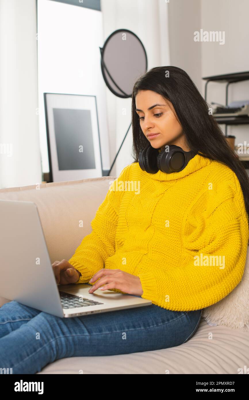 Young Multiracial Woman a yellow sweater working Modern Laptop at the video conference a cozy sofa at home, in Wireless Headphones Digital Technology Stock Photo