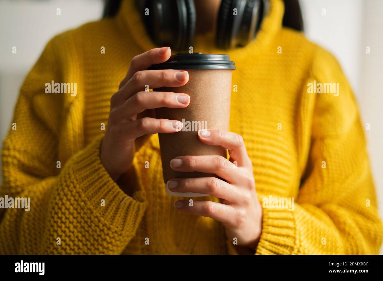 Close-up of an unrecognizable young woman holding recyclable takeaway coffee cup in a yellow sweater on headphones standing by the window at home Stock Photo