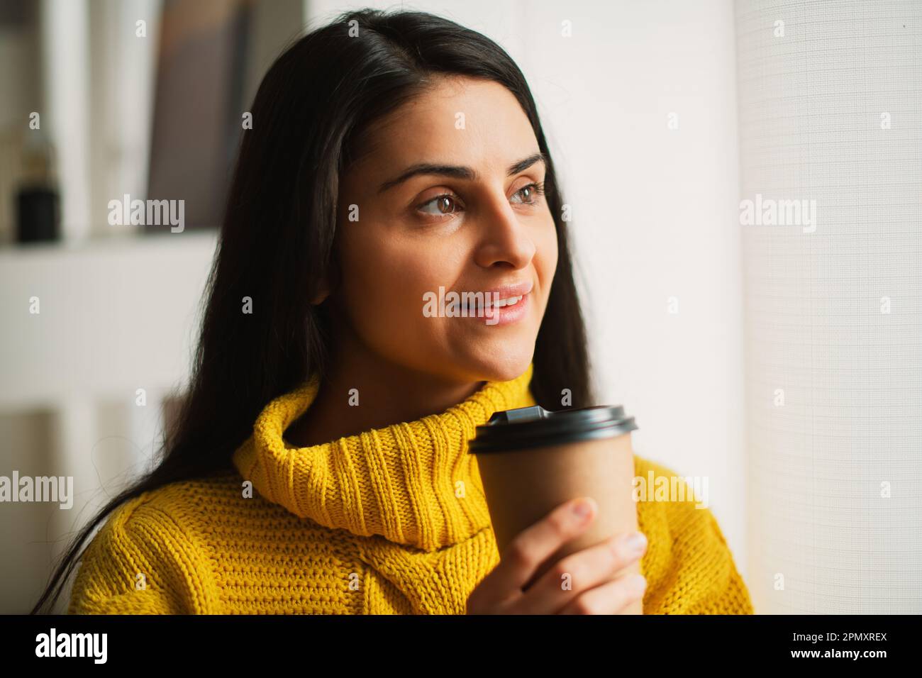 Portrait of smiling happy young multiracial woman's hands holding recycable takeaway coffee cup in a yellow sweater, standing by the window at home Stock Photo