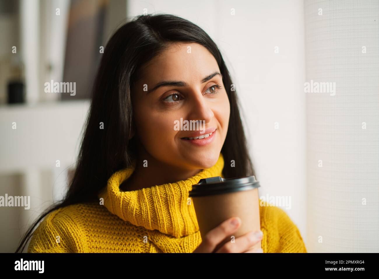 Portrait of smiling happy young multiracial woman's hands holding recycable takeaway coffee cup in a yellow sweater, standing by the window at home Stock Photo
