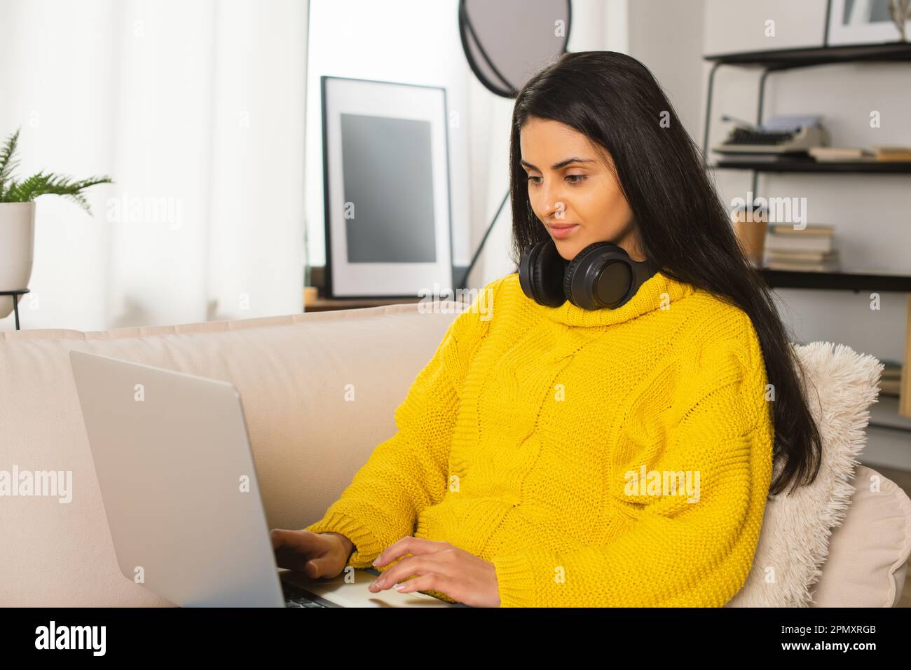 Young Multiracial Woman a yellow sweater working Modern Laptop at the video conference a cozy sofa at home, in Wireless Headphones Digital Technology Stock Photo