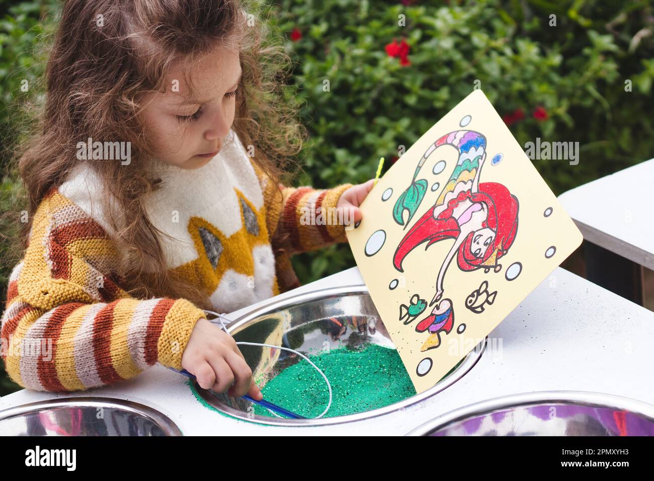 A little girl playing with colored sand to make a cartoon picture of a mermaid Stock Photo