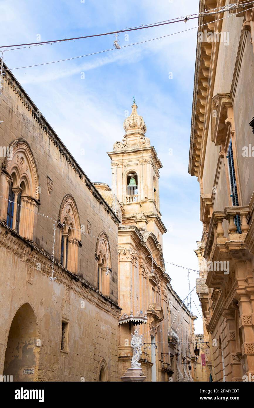 Mdina, Malta - November 13, 2022: Street with limestone buildings and Carmelite Church, also known as Annunciation Church with its belfry and a Virgin Stock Photo