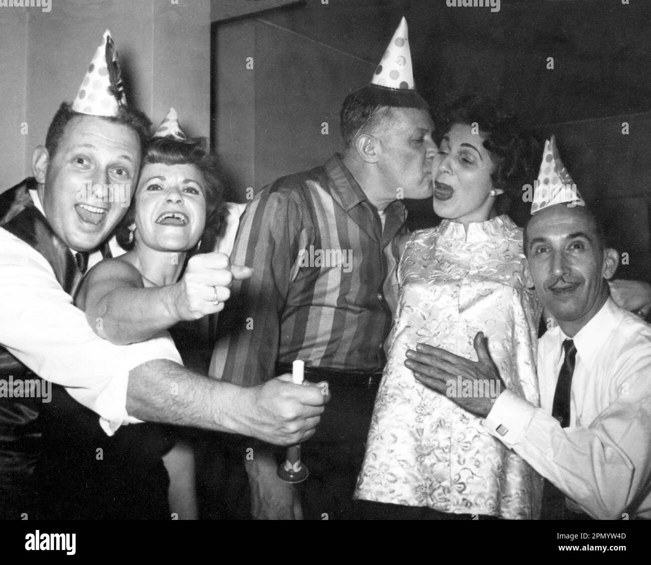 Circa 1960: Adults celebrating at home wearing party hats and holding noisemakers: New Year's Eve USA. 1 woman pregnant. Original black and white vintage photograph. Stock Photo