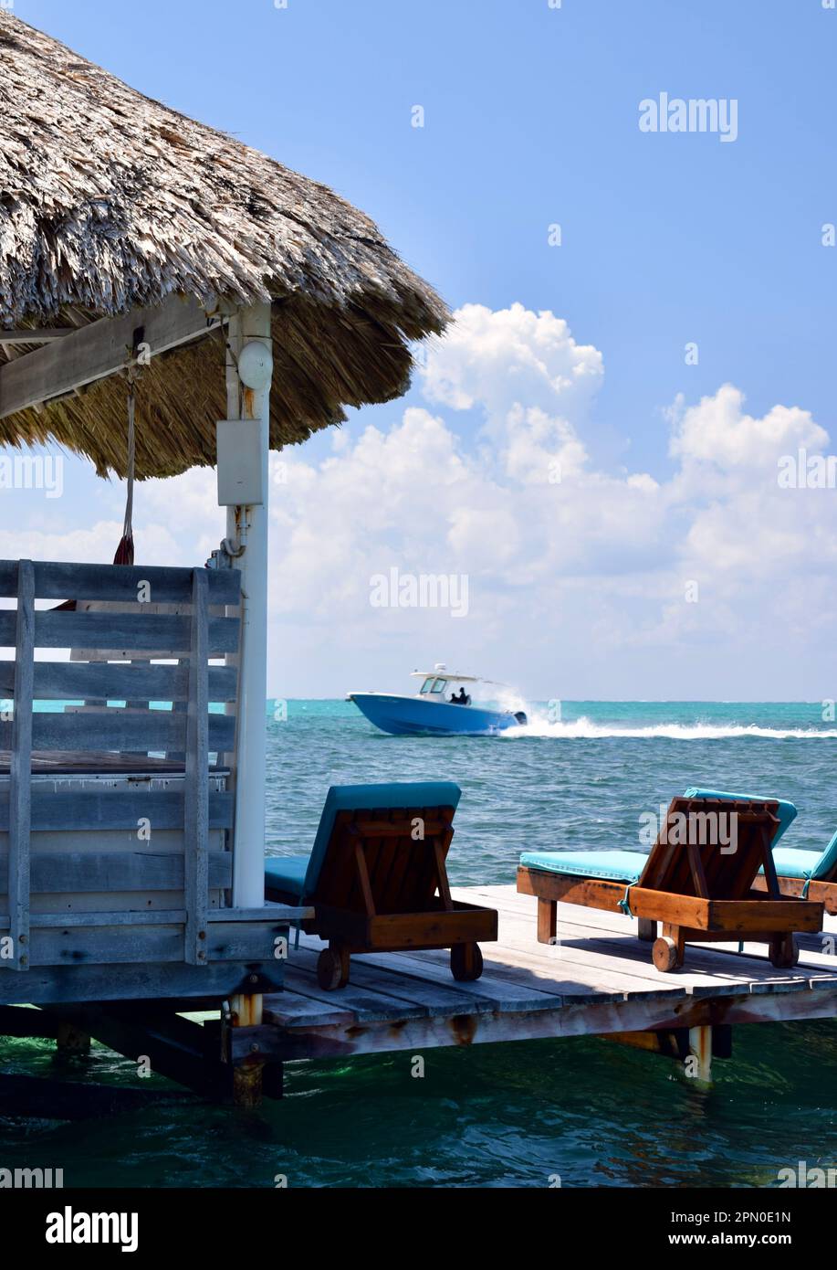 Lounge chairs on a dock and next to a palapa, facing the sea and a passing boat, in San Pedro, Ambergris Caye, Belize, Caribbean/Central America. Stock Photo