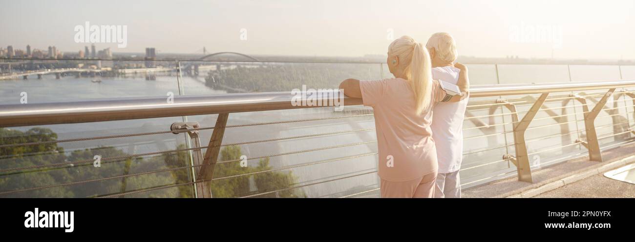 Senior lady hugs husband in sports clothes enjoying city view from footbridge Stock Photo