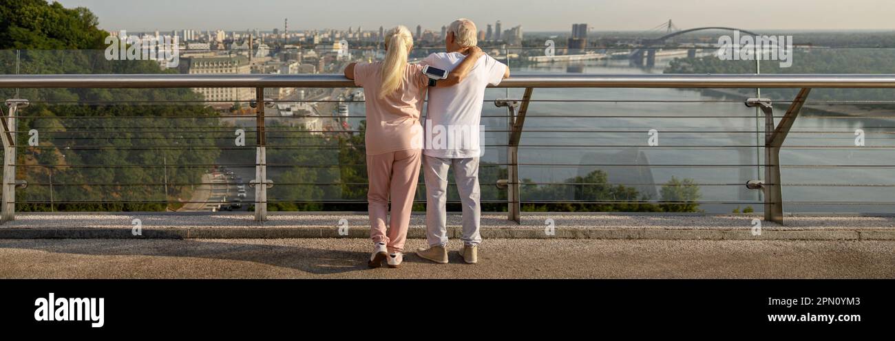 Mature woman hugs man in sportsweat leaning onto railing on contemporary footbridge Stock Photo